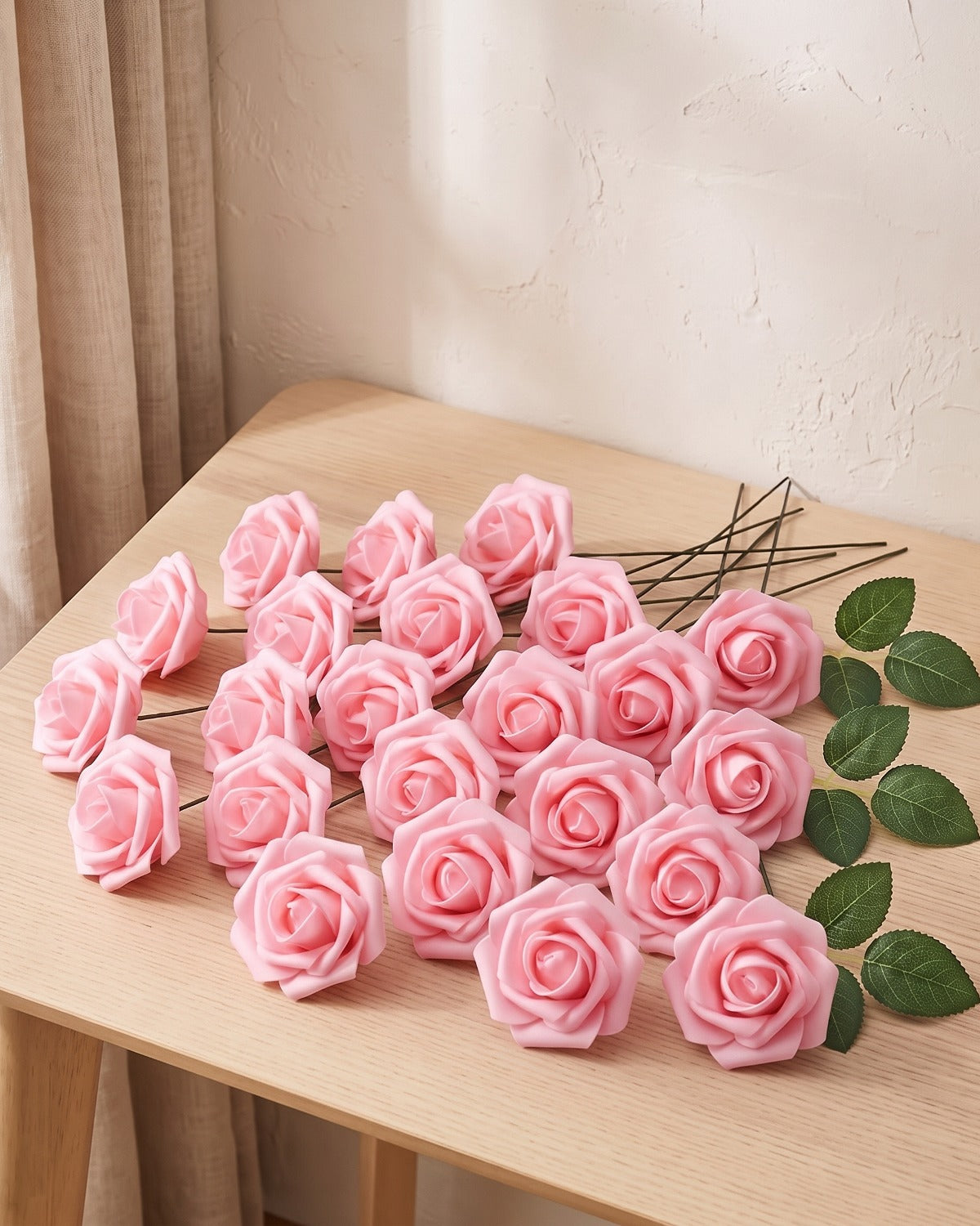 Twenty-five pink foam rose heads on wire stems with green leaf sprays, laid out on a pale oak table