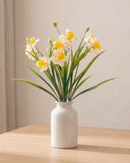 White and yellow artificial daffodil bunch standing in a plain off-white vase on a pale oak table; no vase is included