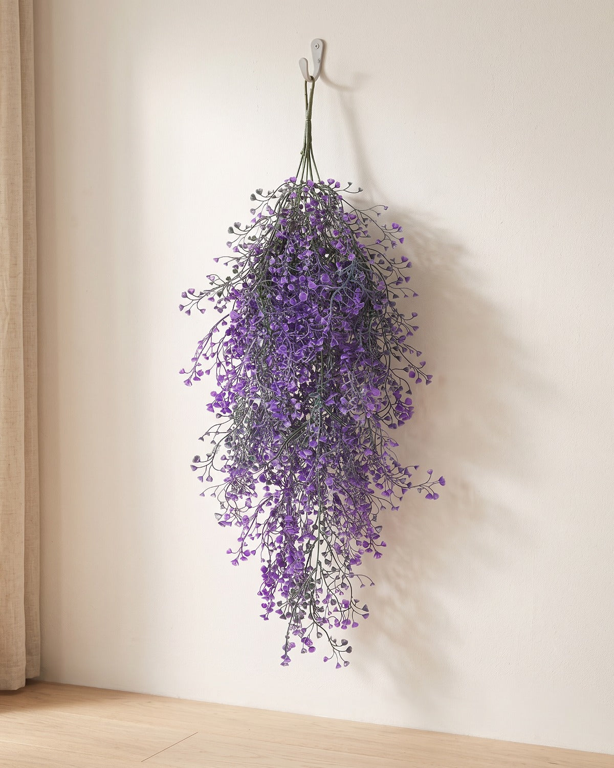 An artificial wisteria spray hanging from a simple hook on a pale wall, its full drop clear of the floor