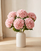 Artificial hydrangea flowers standing in a plain off-white vase on a pale oak table; no vase is included