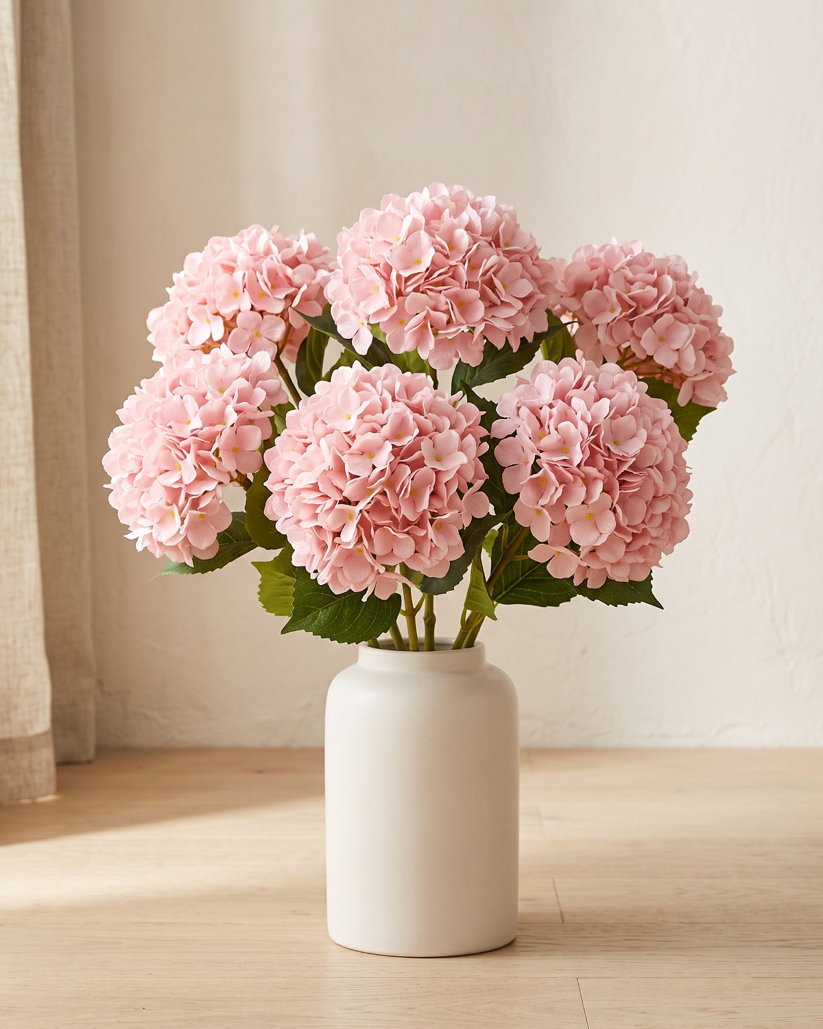 Artificial hydrangea flowers standing in a plain off-white vase on a pale oak table; no vase is included