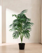 An artificial palm leaf bush with eighteen fronds in a plain black nursery pot on pale oak floorboards in a bright room; pot not included