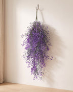 Purple artificial wisteria vine hanging from a simple hook on an off-white plaster wall, its full drop clear of the floor