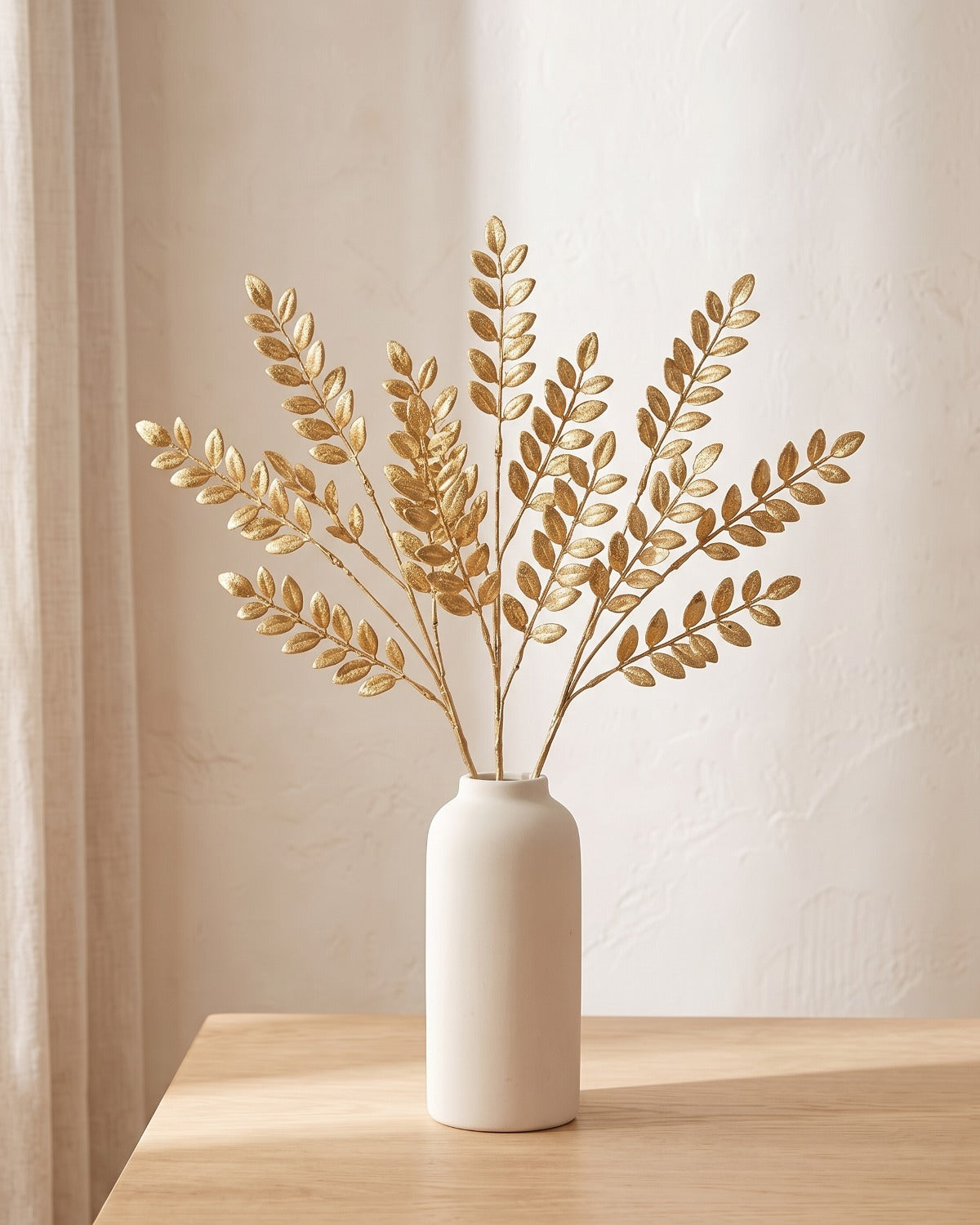 Ten artificial gold leaf stems standing in a plain off-white vase on a pale oak table; no vase is included