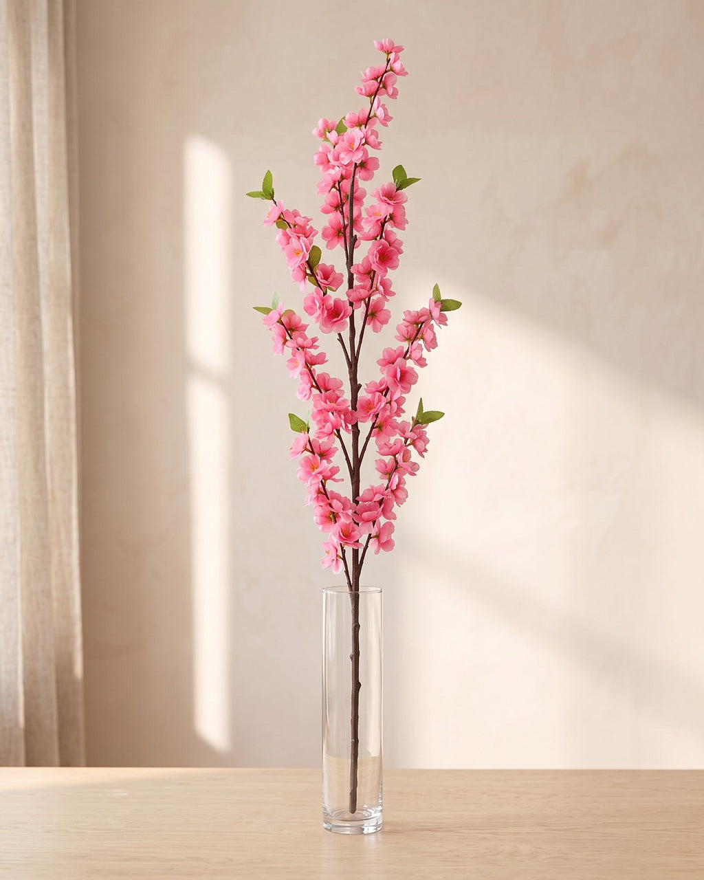 Pink artificial peach blossom branch standing in a tall glass vase on a pale oak table; no vase is included