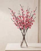Pink artificial plum blossom stems standing in a vase on a pale oak table; no vase is included