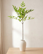 Fine leaf artificial tree branch standing in a plain off-white vase on a pale oak table; no vase is included