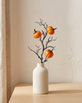 Artificial fall pick, a bare twig carrying small orange pumpkins, in a plain off-white bud vase on a pale oak tabletop, vase not included