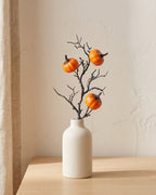 Artificial fall pick, a bare twig carrying small orange pumpkins, in a plain off-white bud vase on a pale oak tabletop, vase not included