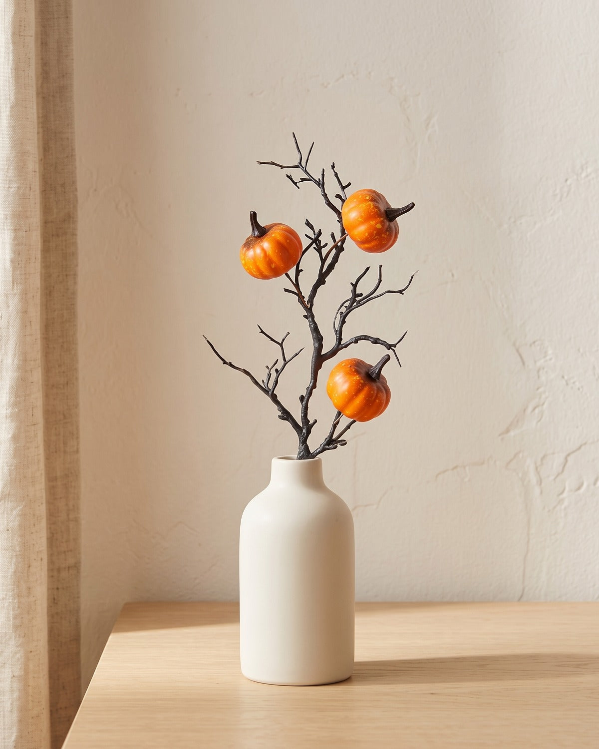 Artificial fall pick, a bare twig carrying small orange pumpkins, in a plain off-white bud vase on a pale oak tabletop, vase not included