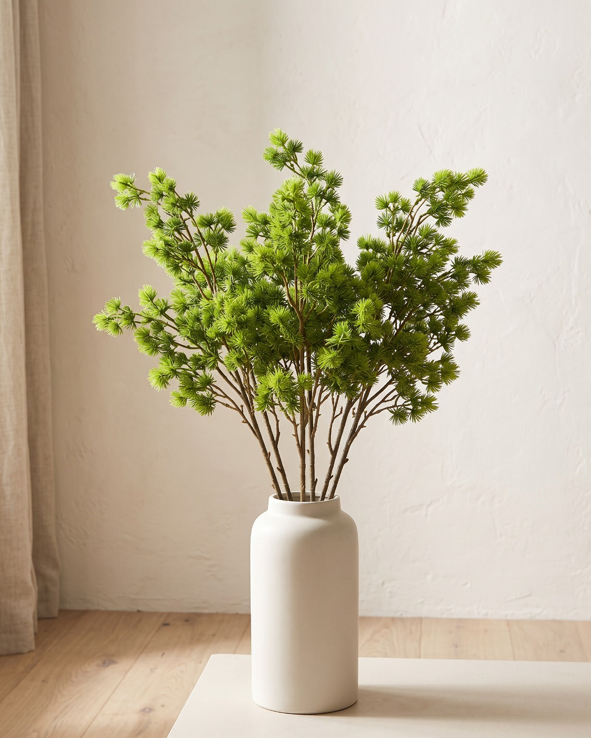 An artificial pine branch standing in a plain off-white vase on a pale oak table; each order is one branch, and no vase is included