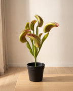 An artificial Venus flytrap in a plain nursery pot on a pale oak table in a bright room; pot not included