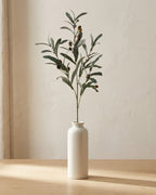 Artificial olive branch in a plain off-white bud vase on a pale oak tabletop, vase not included
