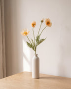A champagne artificial poppy stem, one stem with two open flowers and a bud, in a plain off-white bud vase on a pale oak table; vase not included