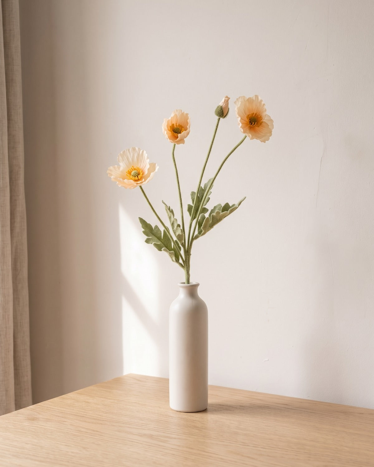 A champagne artificial poppy stem, one stem with two open flowers and a bud, in a plain off-white bud vase on a pale oak table; vase not included