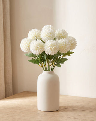 Artificial pompom chrysanthemums standing in a plain off-white vase on a pale oak table; no vase is included