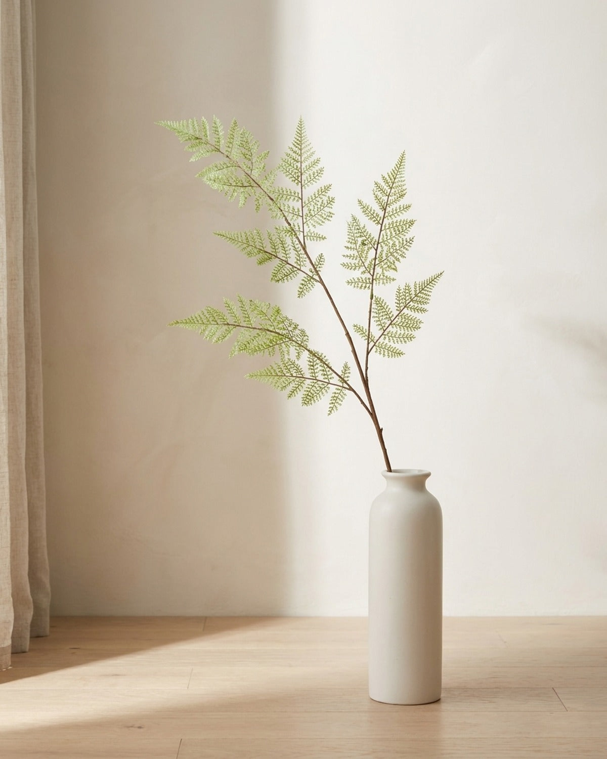 A single artificial fern branch in a plain off-white floor vase on pale oak floorboards, vase not included
