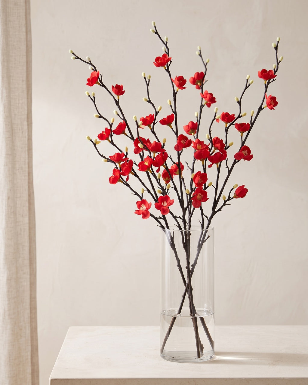 Red artificial plum blossom stems standing in a vase on a pale oak table; no vase is included