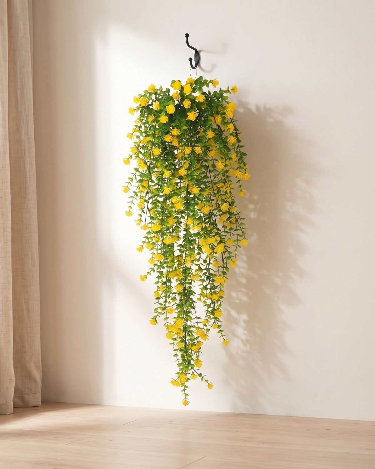 One artificial flowering hanging vine with yellow blossoms, on a black iron hook against a warm plaster wall; hook not included