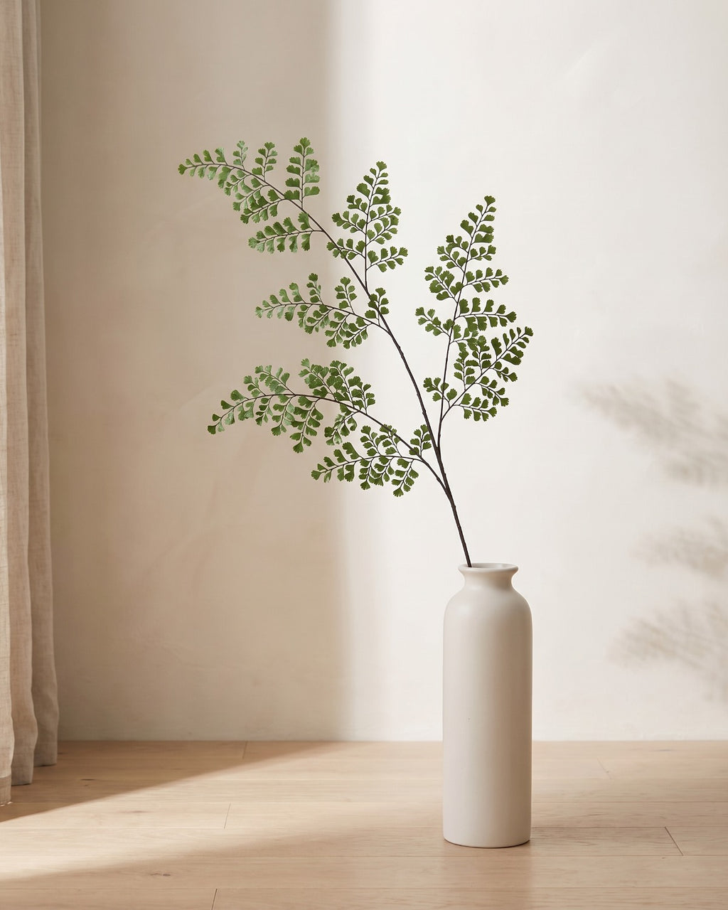 A single artificial maidenhair fern frond, small rounded leaflets on wiry stems, in a plain off-white floor vase on pale oak floorboards (vase not included).