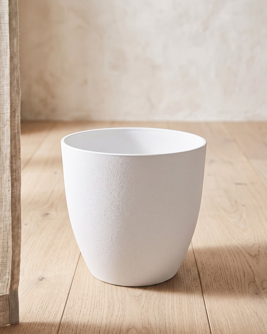 White stone effect plant pot standing empty on pale oak floorboards against an off-white plaster wall