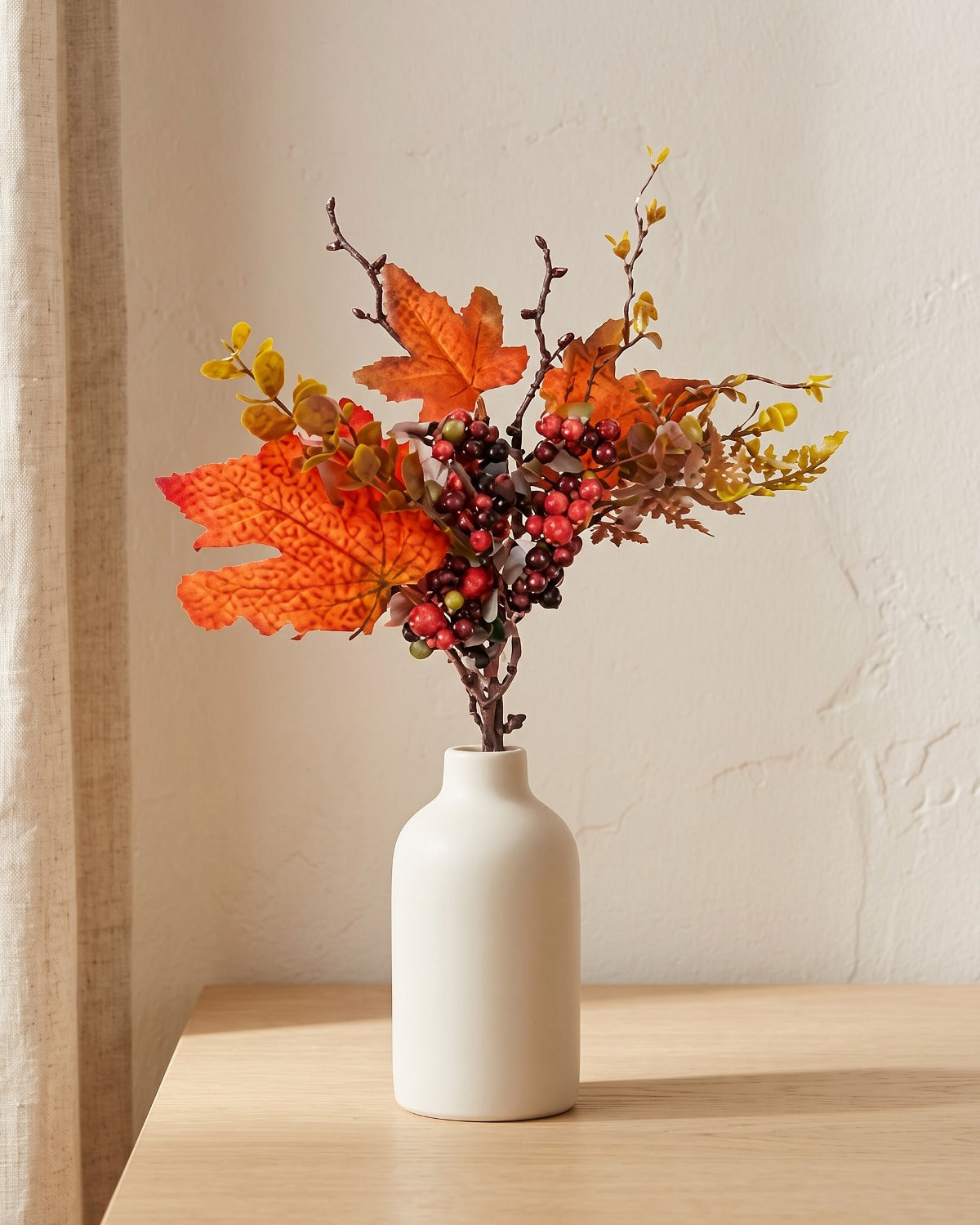 Artificial fall pick of red maple leaves and dark berries, in a plain off-white bud vase on a pale oak tabletop, vase not included