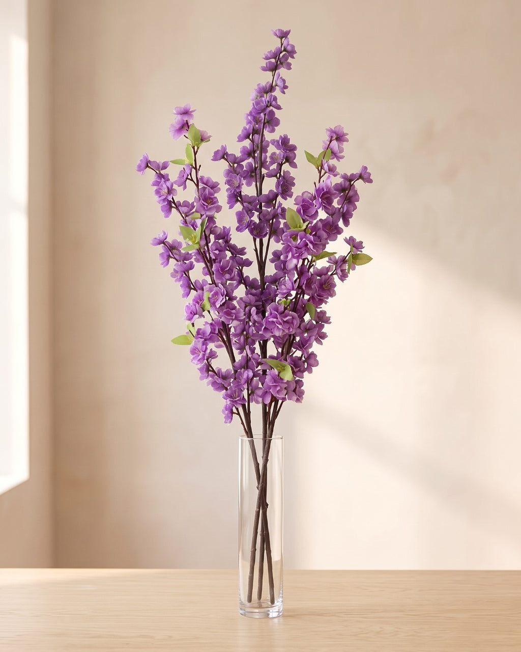 Purple artificial peach blossom branch standing in a tall glass vase on a pale oak table; no vase is included