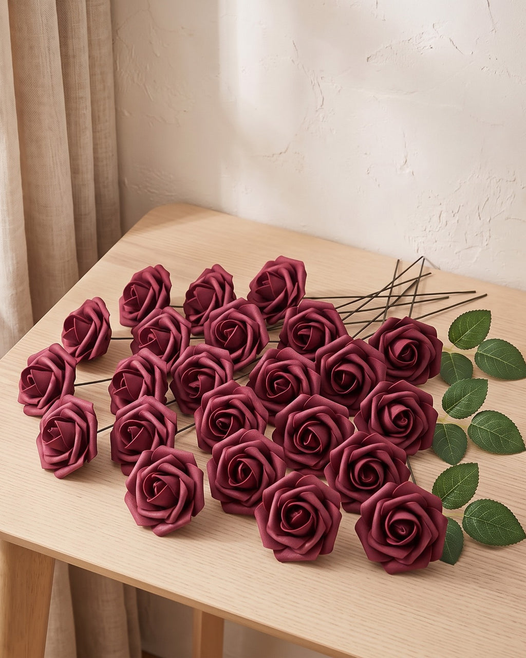 Twenty-five burgundy foam rose heads on wire stems with green leaf sprays, laid out on a pale oak table