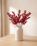 Five artificial red berry branches in a plain off-white vase on a pale oak table; vase not included