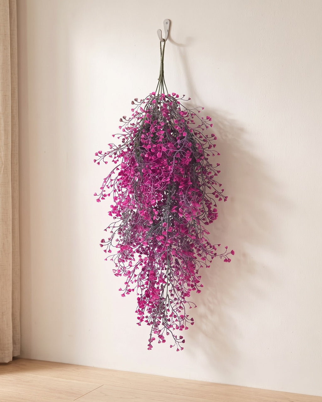 Magenta artificial wisteria vine hanging from a simple hook on an off-white plaster wall, its full drop clear of the floor