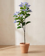 Artificial variegated leaf branch with lilac leaf tips, in a plain terracotta pot against a warm plaster wall; pot not included