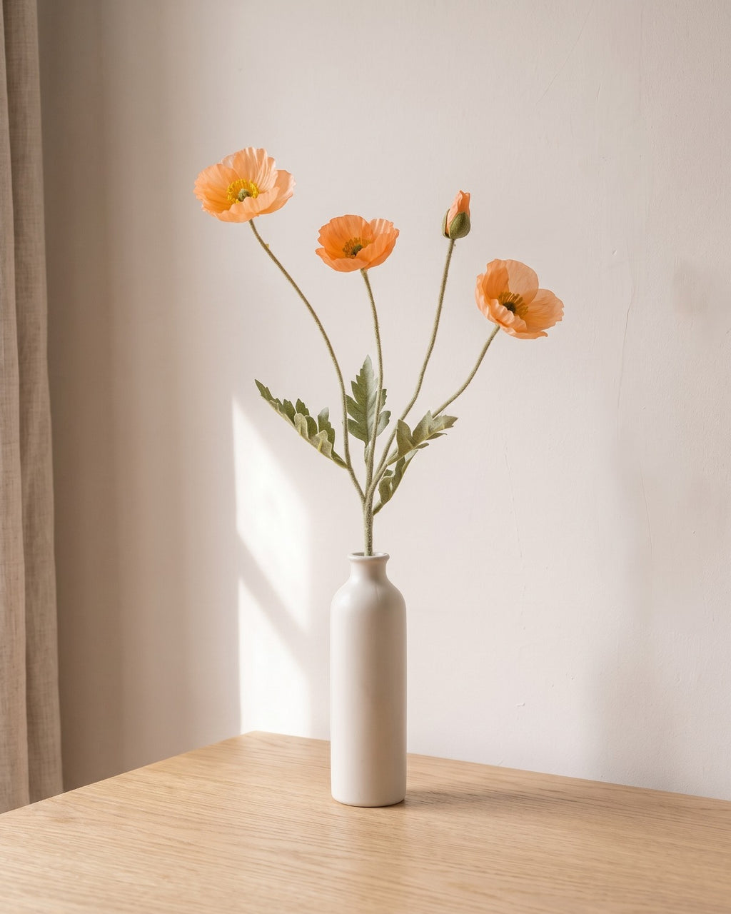 A mellow apricot artificial poppy stem, one stem with two open flowers and a bud, in a plain off-white bud vase on a pale oak table; vase not included
