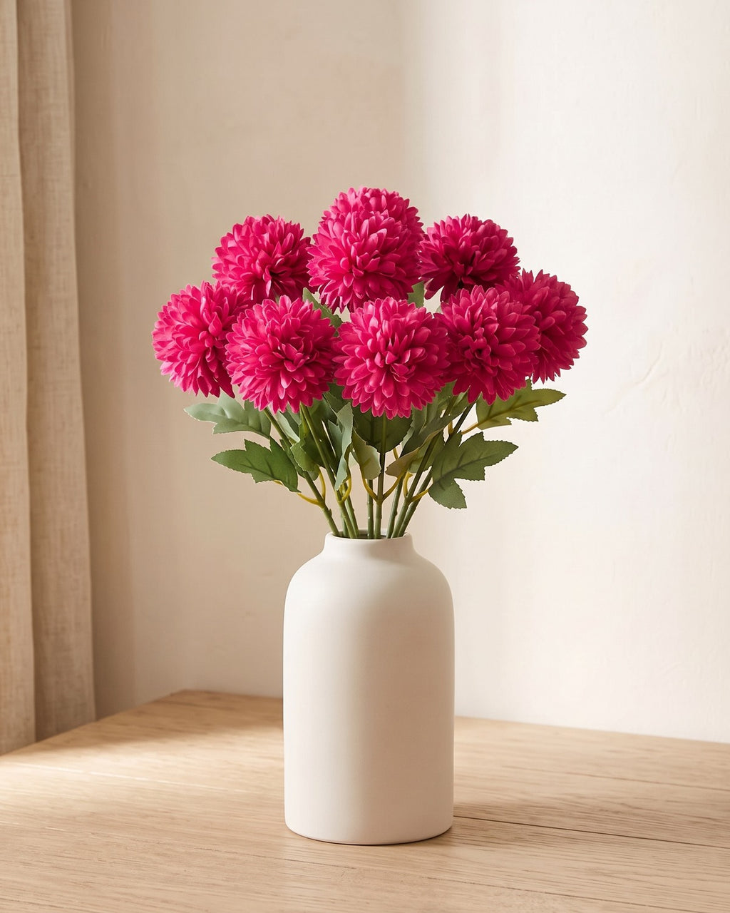 Magenta artificial pompom chrysanthemums standing in a vase on a pale oak table; no vase is included