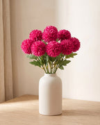 Magenta artificial pompom chrysanthemums standing in a vase on a pale oak table; no vase is included