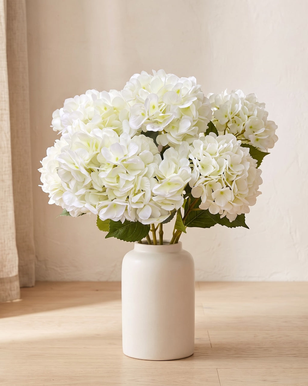 Milk White artificial hydrangea flowers standing in a plain off-white vase on a pale oak table; no vase is included