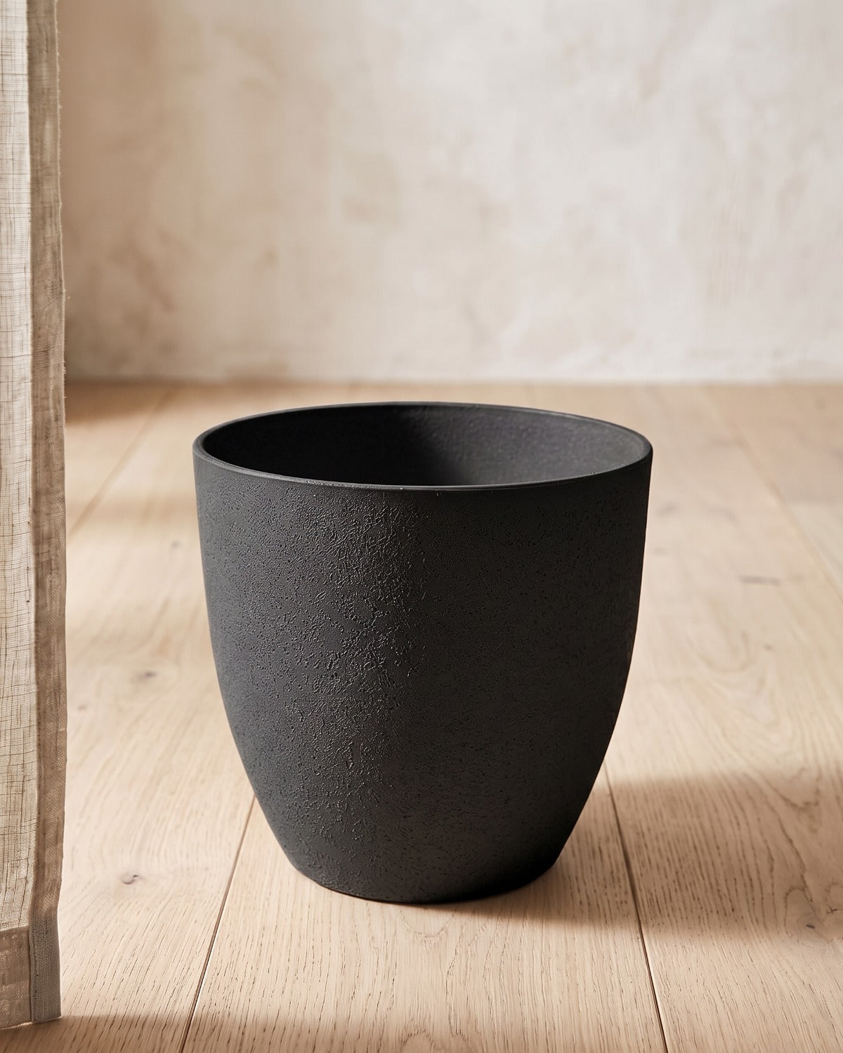 Black stone effect plant pot standing empty on pale oak floorboards against an off-white plaster wall