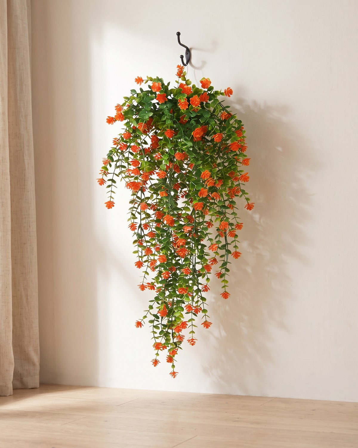 One artificial flowering hanging vine with orange blossoms, on a black iron hook against a warm plaster wall; hook not included