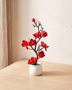 A small artificial plum blossom bonsai in deep rose red bloom, in the white ceramic pot it comes in, on a pale oak table