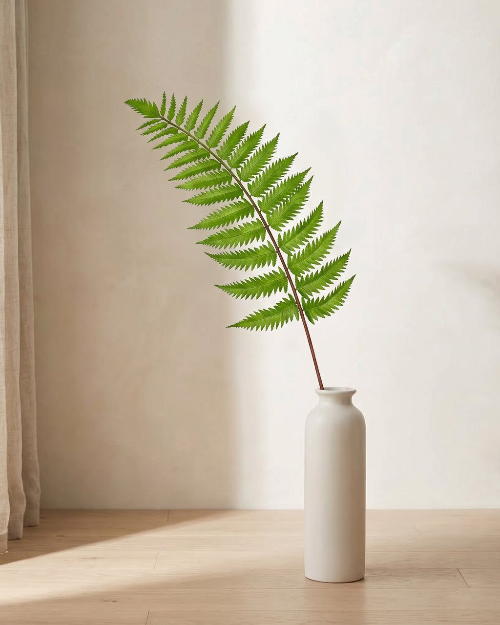 A single artificial Boston fern frond, a broad flat blade of paired leaflets, in a plain off-white floor vase on pale oak floorboards (vase not included).