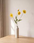 A yellow artificial poppy stem, one stem with two open flowers and a bud, in a plain off-white bud vase on a pale oak table; vase not included