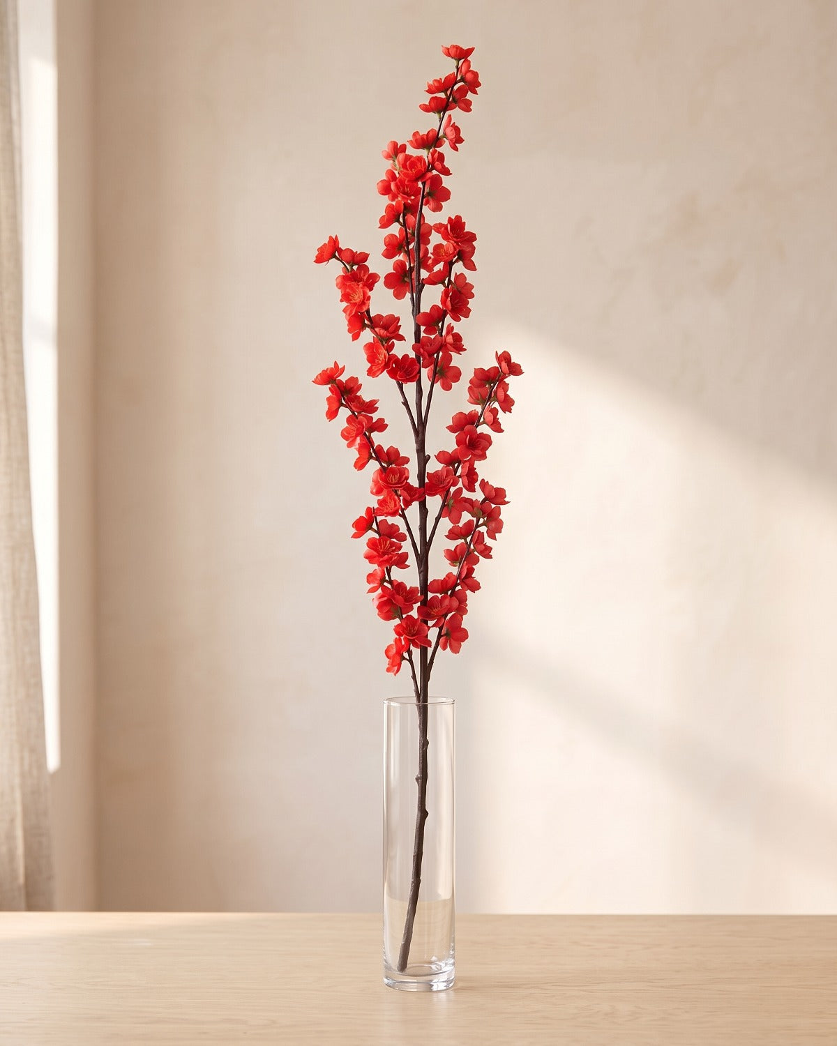 Red artificial peach blossom branch standing in a tall glass vase on a pale oak table; no vase is included