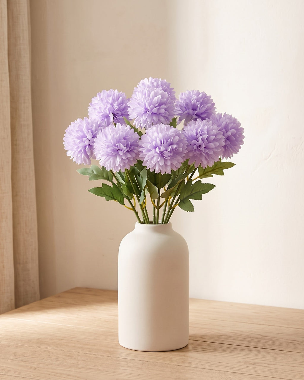 Lilac artificial pompom chrysanthemums standing in a vase on a pale oak table; no vase is included