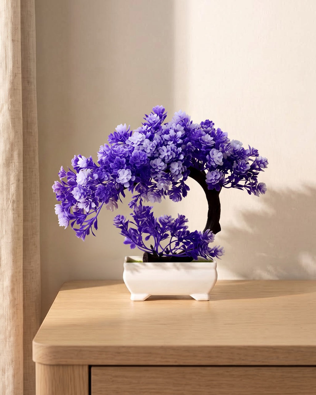 A small artificial bonsai in purple blossom, in the white ceramic tray it comes in, on a pale oak table