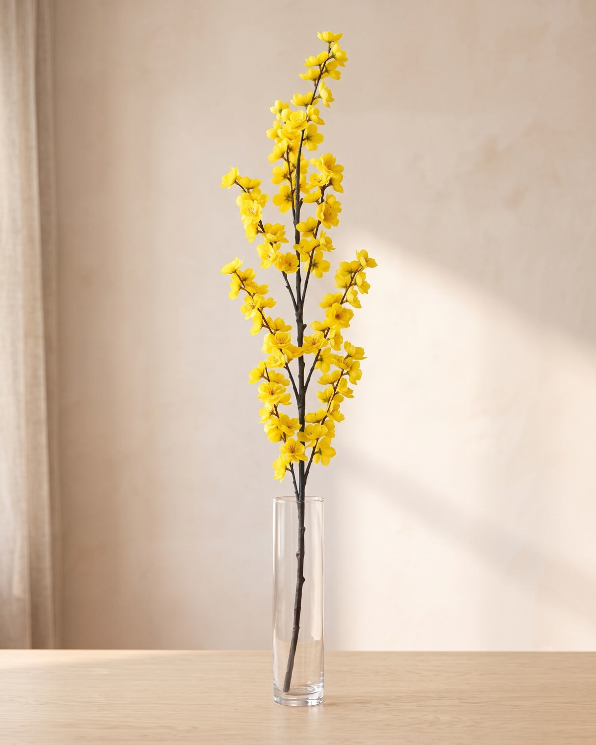 Yellow artificial peach blossom branch standing in a tall glass vase on a pale oak table; no vase is included