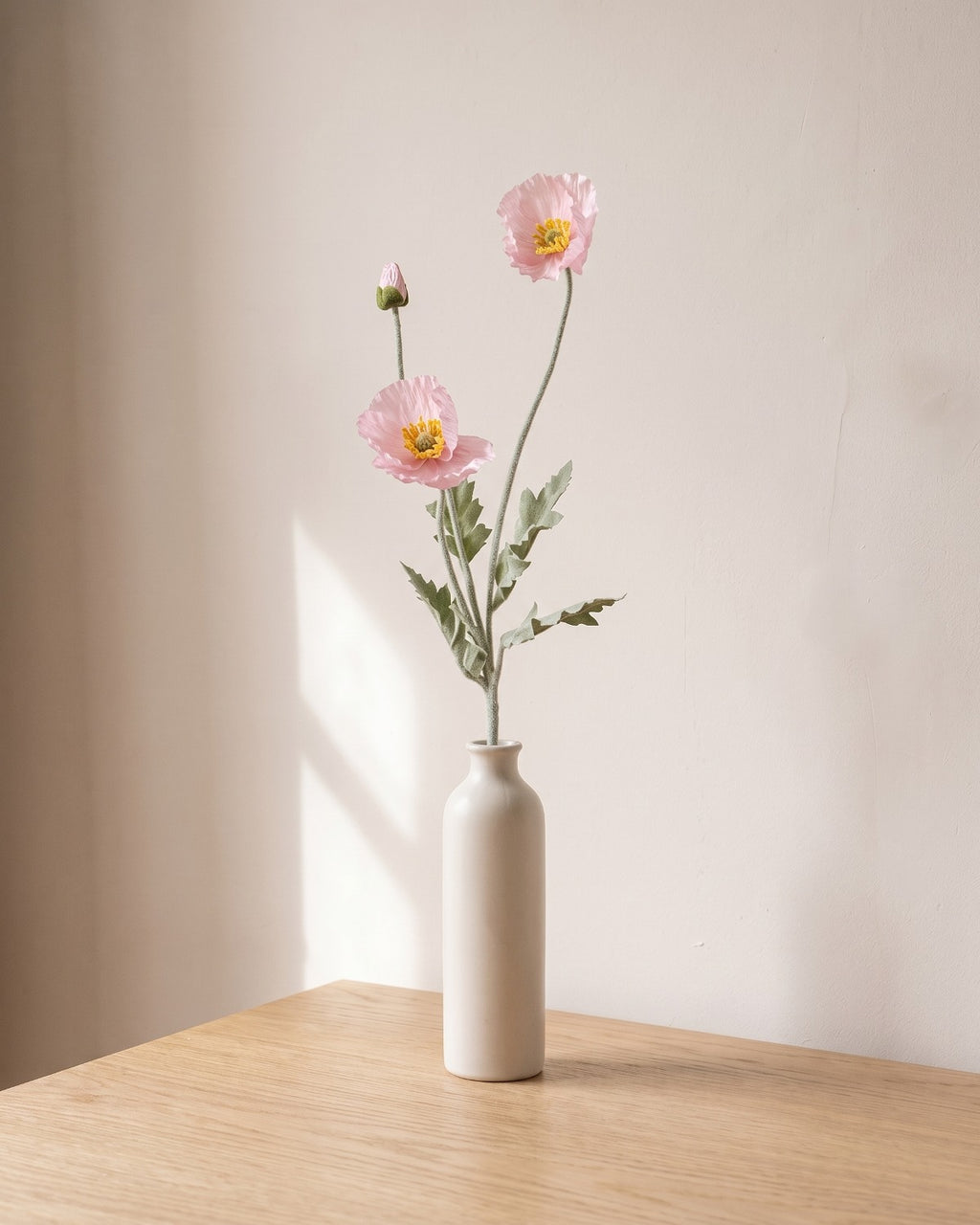 A light pink artificial poppy stem, one stem with two open flowers and a bud, in a plain off-white bud vase on a pale oak table; vase not included