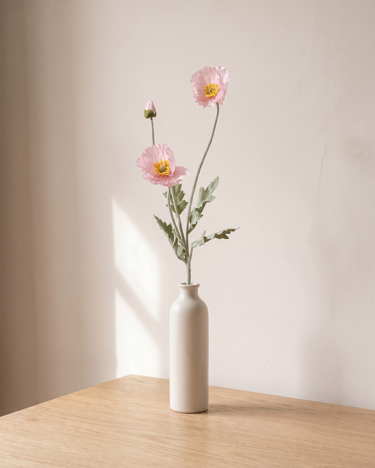 A light pink artificial poppy stem, one stem with two open flowers and a bud, in a plain off-white bud vase on a pale oak table; vase not included