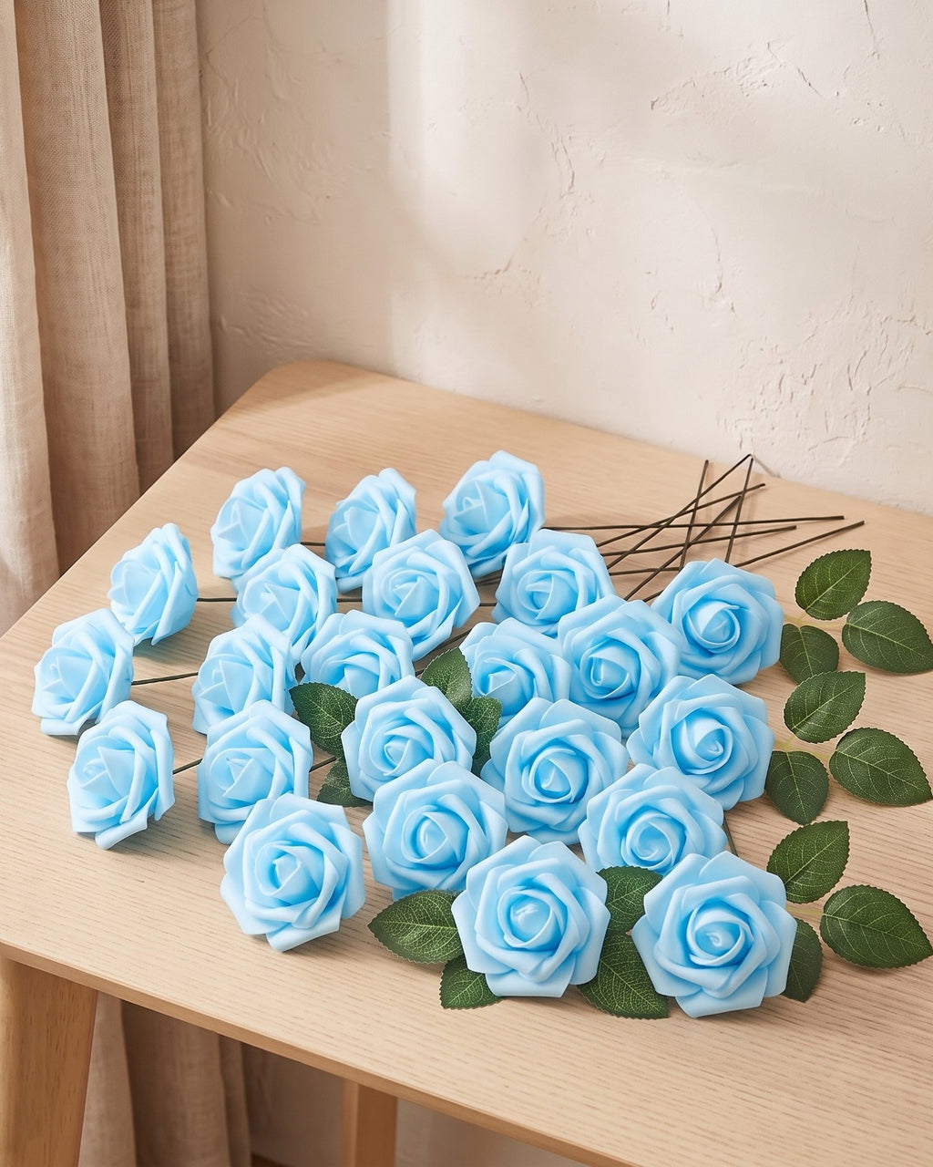 Twenty-five sky blue foam rose heads on wire stems with green leaf sprays, laid out on a pale oak table