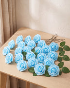 Twenty-five sky blue foam rose heads on wire stems with green leaf sprays, laid out on a pale oak table