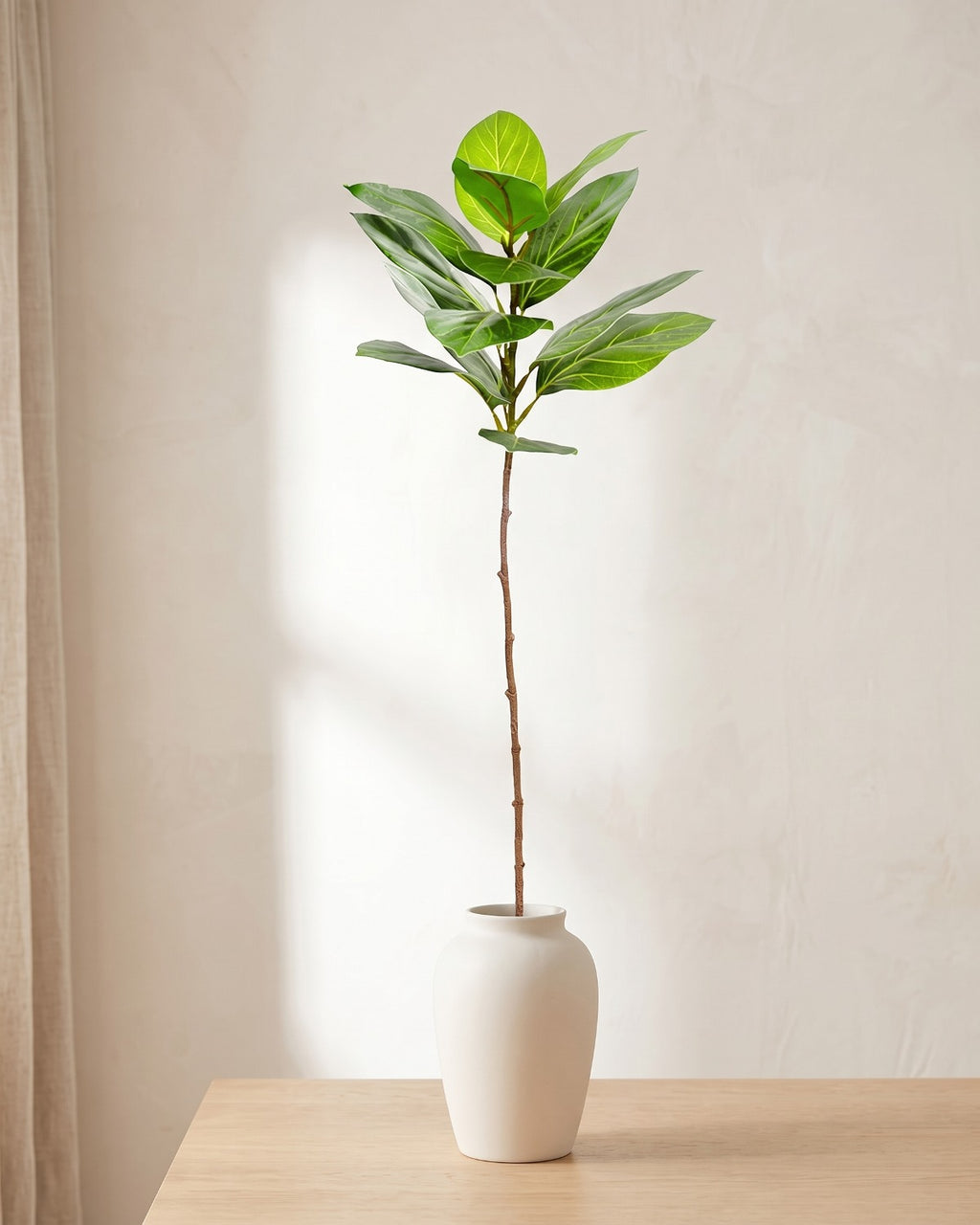 Veined oval leaf artificial tree branch standing in a plain off-white vase on a pale oak table; no vase is included