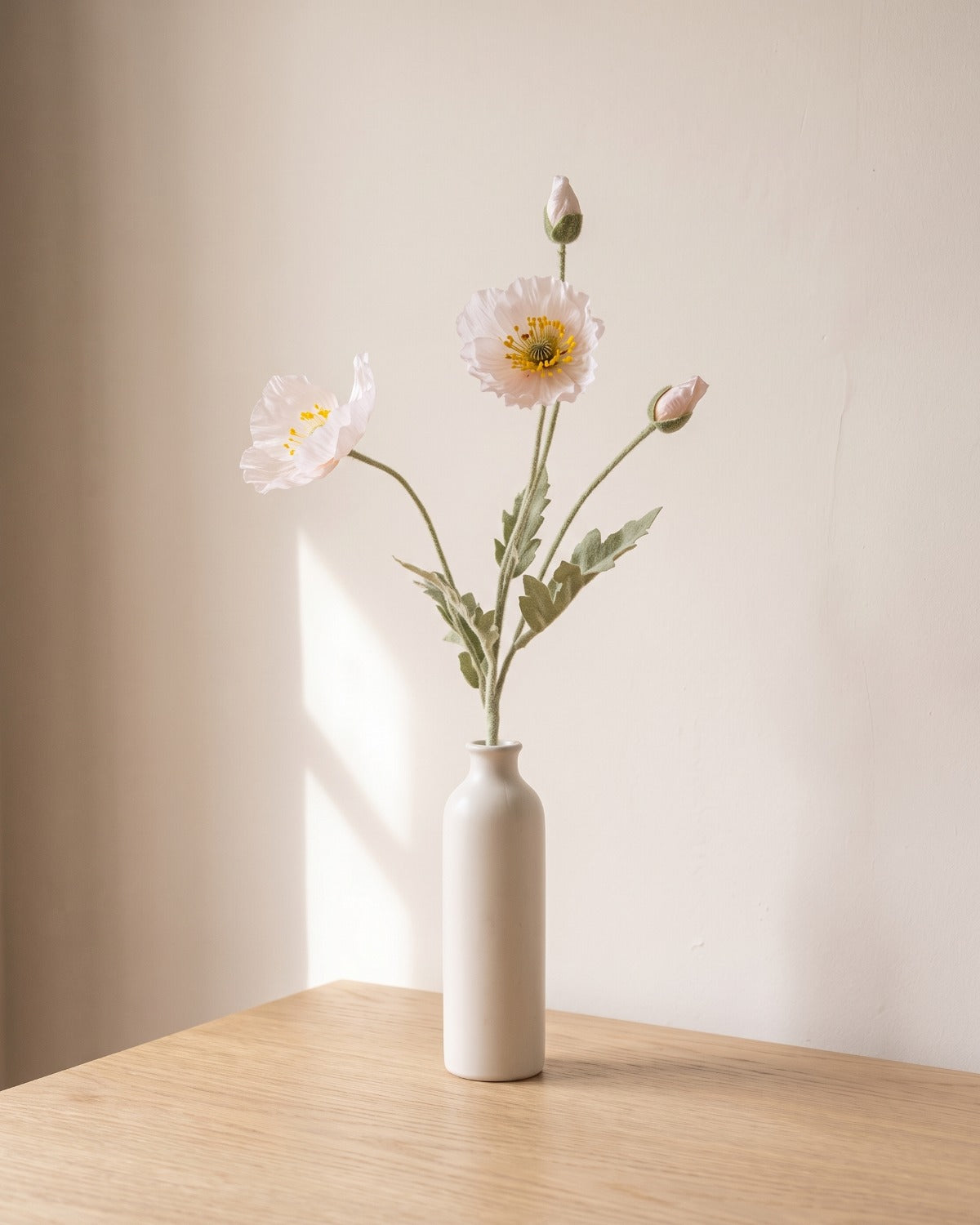 A ice pink artificial poppy stem, one stem with two open flowers and a bud, in a plain off-white bud vase on a pale oak table; vase not included